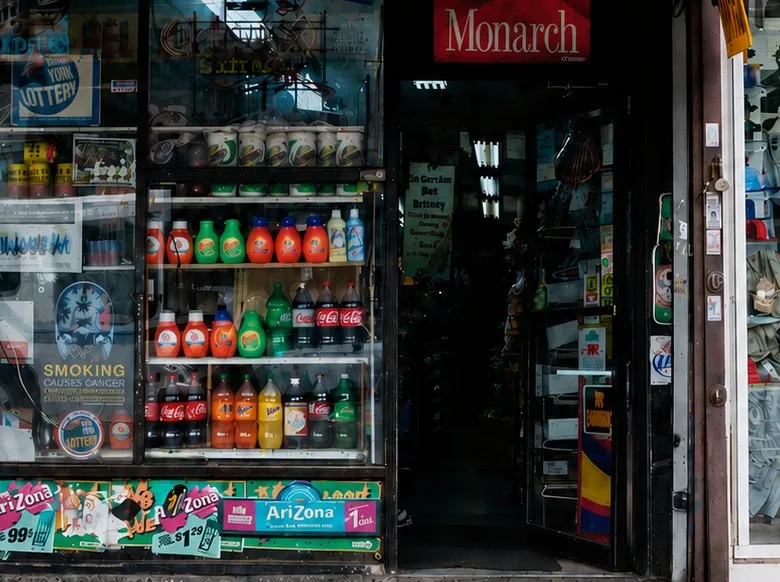 The El Bloque window: a refrigerator case packed with soda and cleaning products behind glass carrying lottery signs and AriZona price decals